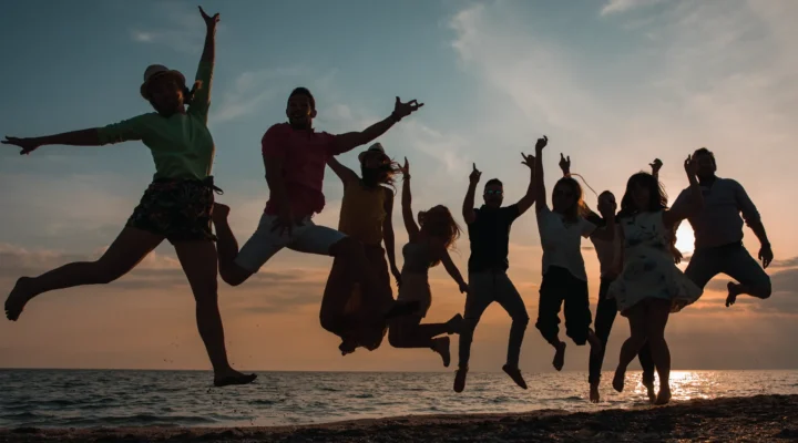 Silhouettes of a group of happy people jumping in the air on a beach during sunset, celebrating together. A red graphic overlay on the left reads: 'WHY EMPLOYEE RECOGNITION DRIVES ENGAGEMENT AND RETENTION #WORKPLACECULTURE'.