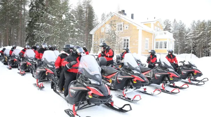 A group of corporate participants in red and black winter gear sitting on a line of Lynx snowmobiles in front of a yellow wooden house in a snowy forest.
